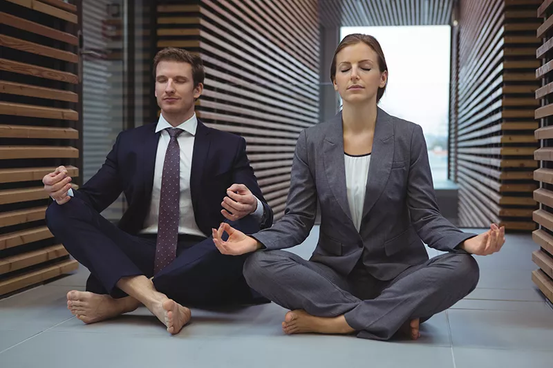 Dieses Bild zeigt einen Mann und eine Frau, sitzend beim Yoga in Businesskleidung